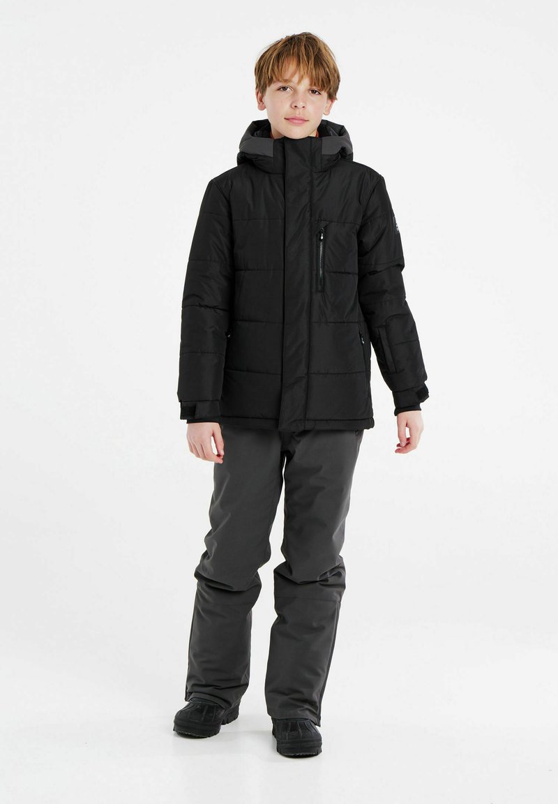 Schwarze gepolsterte Jacke mit hohem Kragen und Kapuze, ausgestattet mit Reißverschluss- und Klappentaschen. Kombiniert mit grauen isolierten Hosen und schwarzen Stiefeln.