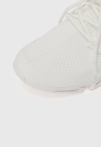 Zapatilla deportiva blanca con un upper texturizado, que presenta una puntera redondeada y cordones. La suela es ligeramente translúcida con un diseño curvado.