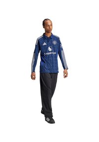 Blauw sportjersey met witte details, voorzien van een logo en merknaam. Gekleed met zwarte broek en schoenen. Textuur lijkt glad en ademend.