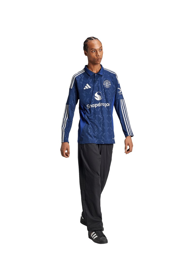 Blauw sportjersey met witte details, voorzien van een logo en merknaam. Gekleed met zwarte broek en schoenen. Textuur lijkt glad en ademend.