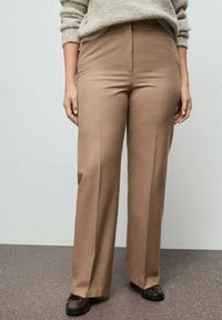 Pantalones marrones de pierna ancha hechos de una tela suave con una cintura alta, que presentan pliegues frontales, combinados con zapatos oscuros y un suéter ligero.