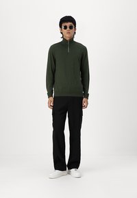 Sweater vert zippé à manches longues, associé à un pantalon cargo noir. Le modèle porte des lunettes de soleil rondes et des baskets blanches.