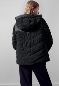 Giacca puffer nera in corduroy con cappuccio, caratterizzata da un design trapuntato e un pannello a contrasto sulle spalle. Texture morbida e vestibilità comoda.