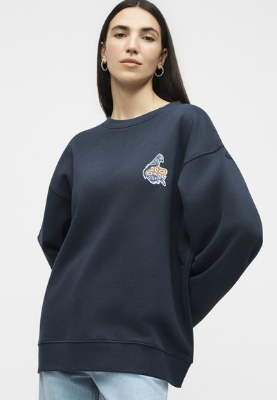 Femme portant un sweat-shirt bleu marine avec un oiseau brodé et un signe dollar sur la poitrine, associé à un jean bleu clair et des boucles d'oreilles anneaux.