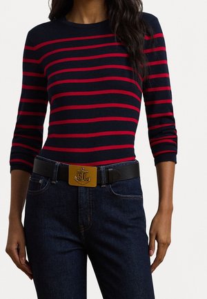 Kvinde iført navyblå og rødstribet langærmet top, mørkeblå jeans og et sort bælte med en guldbeslag med et anker og initialer.