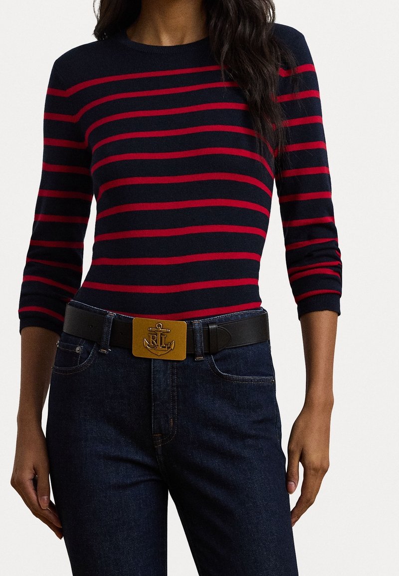 Femme portant un haut à manches longues rayé bleu marine et rouge, un jean bleu foncé, et une ceinture noire avec une boucle dorée ornée d'une ancre et d'initiales.