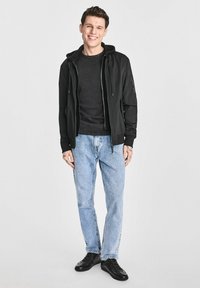 Czarna bluza z kapturem na zamek rozpięta na ciemnoszarym T-shircie, jasnoniebieskie dżinsy i czarne trampki. Model ma krótkie włosy i stoi na jednolitym tle.