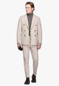 Completo beige a righe con giacca doppiopetto e pantaloni slim-fit, abbinato a un dolcevita scuro e scarpe di pelle marroni.
