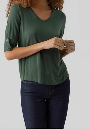 Mujer con camisa de cuello en V verde oscuro con mangas remangadas y vaqueros azul oscuro, de pie frente a un fondo liso de color gris claro.