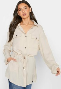 Beige Bluse mit Knöpfen, langen Ärmeln, Brusttaschen und tailliertem Schnitt. Glattes Material mit lässiger Passform, verziert mit silberenen Hardware-Details.