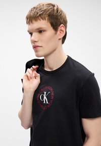 Czarny bawełniany t-shirt z okrągłym logo z napisem "CALVIN KLEIN NEW YORK 1968" w czerwonym kolorze wokół dużego białego "CK" na środku.
