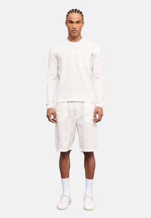 Uomo con i capelli intrecciati in piedi rivolto in avanti, indossa una camicia bianca a maniche lunghe, pantaloncini bianchi fino al ginocchio, calzini bianchi e scarpe da ginnastica bianche.