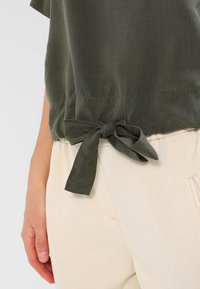 Olive green shirt with a tied knot at the hem, made of smooth fabric. Paired with cream-colored pants, showcasing subtle texture and detail.