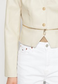 Veste en similicuir crème à coupe ajustée, avec des accessoires dorés, une fermeture éclair et un ourlet raccourci. Associée à un jean blanc.