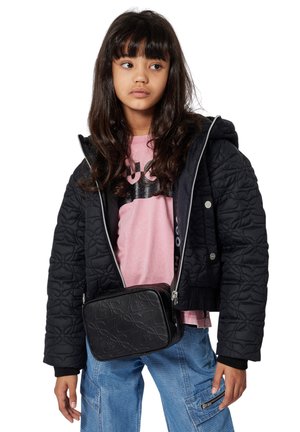 Jeune fille aux longs cheveux bruns, portant une veste matelassée noire, un t-shirt rose, un pantalon bleu et un sac bandoulière noir à motifs.