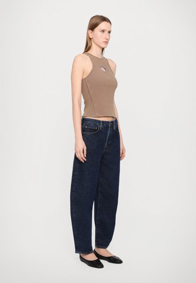 Braun gerippter Tank-Top mit rautenförmigem Ausschnitt, kombiniert mit dunkelblauen Schlagjeans und schwarzen Ballerinas, vor einem schlichten hellen Hintergrund.