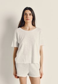 Žena s dlhými hnedými vlasmi, ktorá má na sebe voľný biely krátky rukáv a svetlosivé šortky, stojaca oproti jednoliatemu béžovému pozadiu.