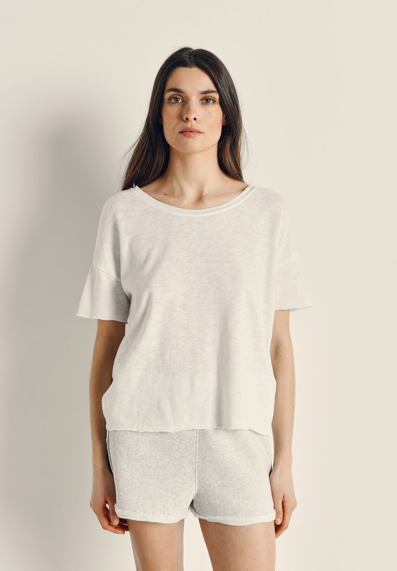 Žena s dlhými hnedými vlasmi, ktorá má na sebe voľný biely krátky rukáv a svetlosivé šortky, stojaca oproti jednoliatemu béžovému pozadiu.