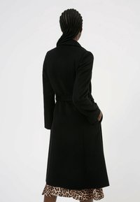 Manteau en mélange de laine noir avec une silhouette longue, taille ceinturée et poches latérales. Comprend un col classique et un tissu texturé.
