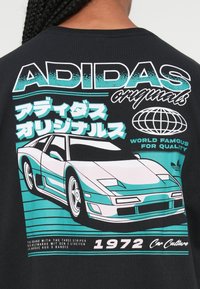 Juodas marškinėlių nugarėlė, rodanti žydros ir baltos spalvos sportinio automobilio grafiką su užrašu "ADIDAS originals", japonišku tekstu ir šūkiu "1972 Car Culture".
