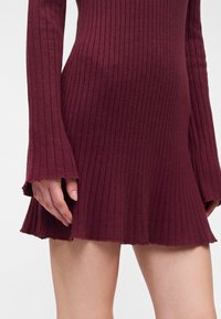 Femme portant une robe en maille ajustée, côtelée, à manches longues, de couleur rouge foncé avec un ourlet évasé, montrant la main et le haut de la cuisse sur un fond blanc.