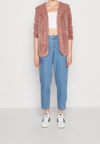 Suddig rosa kardigan med huva, bärs över en vit cropped topp; ihop med lösa blå jeans och vita sneakers med svarta ränder.