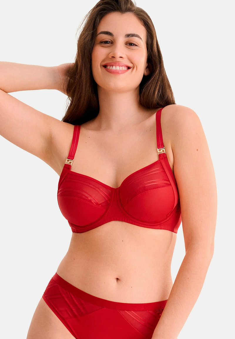 Ensemble de lingerie rouge comprenant un soutien-gorge transparent avec armature, bonnets moulés, bretelles réglables et accessoires en métal doré. Les culottes assorties complètent le design.