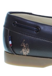 Tummanruskea nahkakenkä, jossa on vaaleanruskea pohja. Mukana on pieni punavalko-sininen lappu ja kohokuvioitu logo, jossa on kaksi polo-pelaajaa.