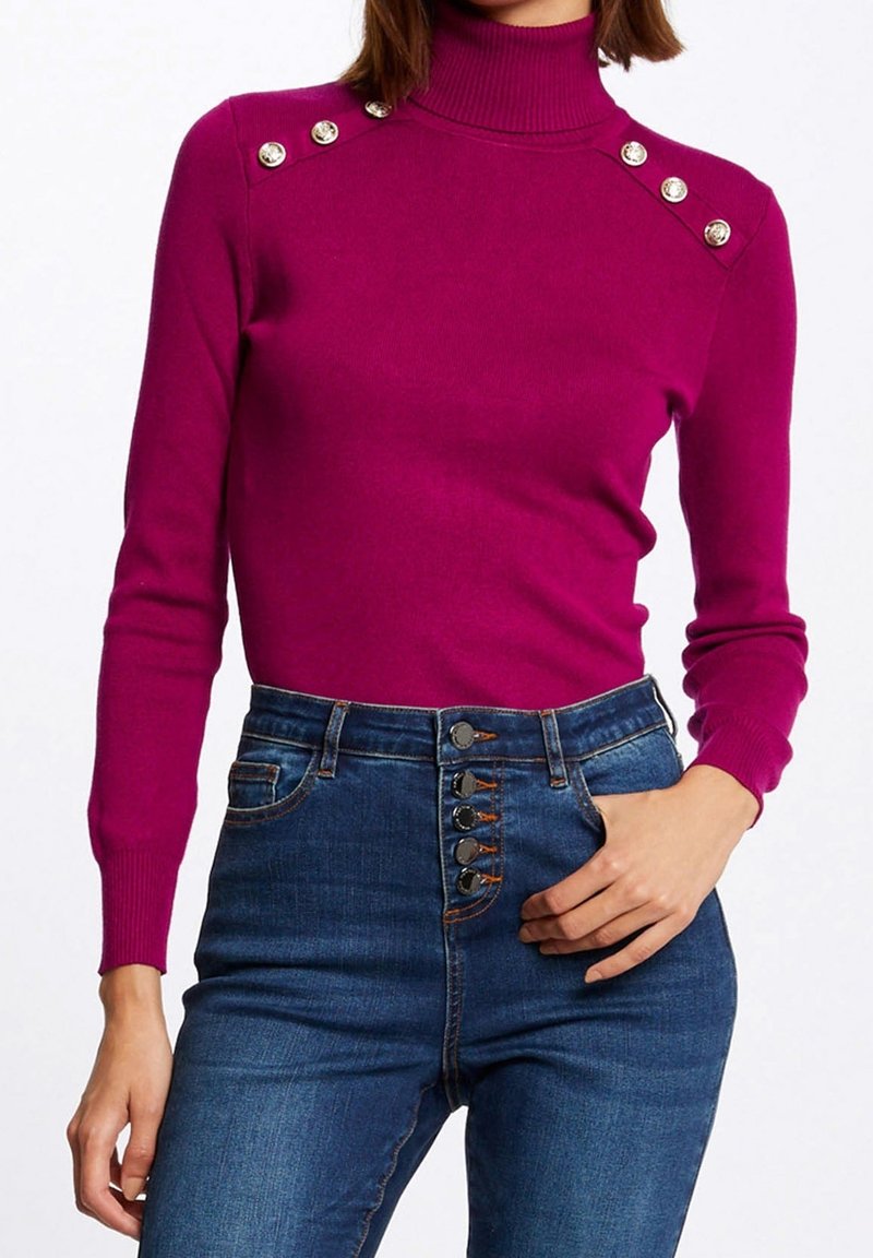 Femme portant un pull col roulé magenta ajusté avec des boutons décoratifs dorés sur les épaules et un jean taille haute bleu avec plusieurs boutons à l'avant.