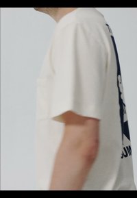 Vue latérale d'une personne portant un T-shirt beige clair avec un grand motif noir et blanc visible dans le dos.