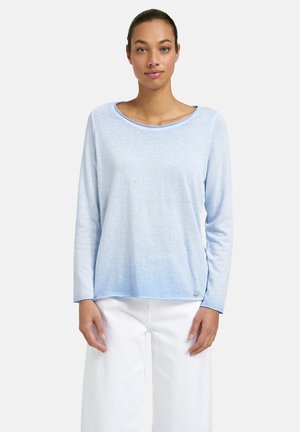 Woman wearing a light blue long-sleeve top and white pants, standing facing forward against a plain white background.