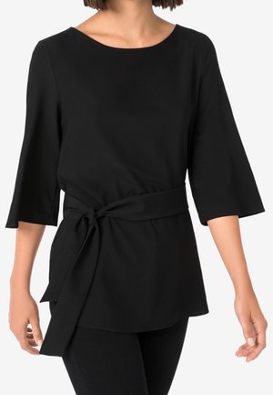 Femme portant un haut noir à manches trois-quarts avec une ceinture nouée à la taille, montrée des épaules aux hanches sur fond blanc.