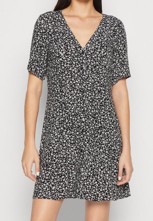 Frau trägt ein kurzes schwarzes Kleid mit weißem Blumenmuster, V-Ausschnitt und kurzen Ärmeln, steht vor einem einfarbigen hellen Hintergrund.