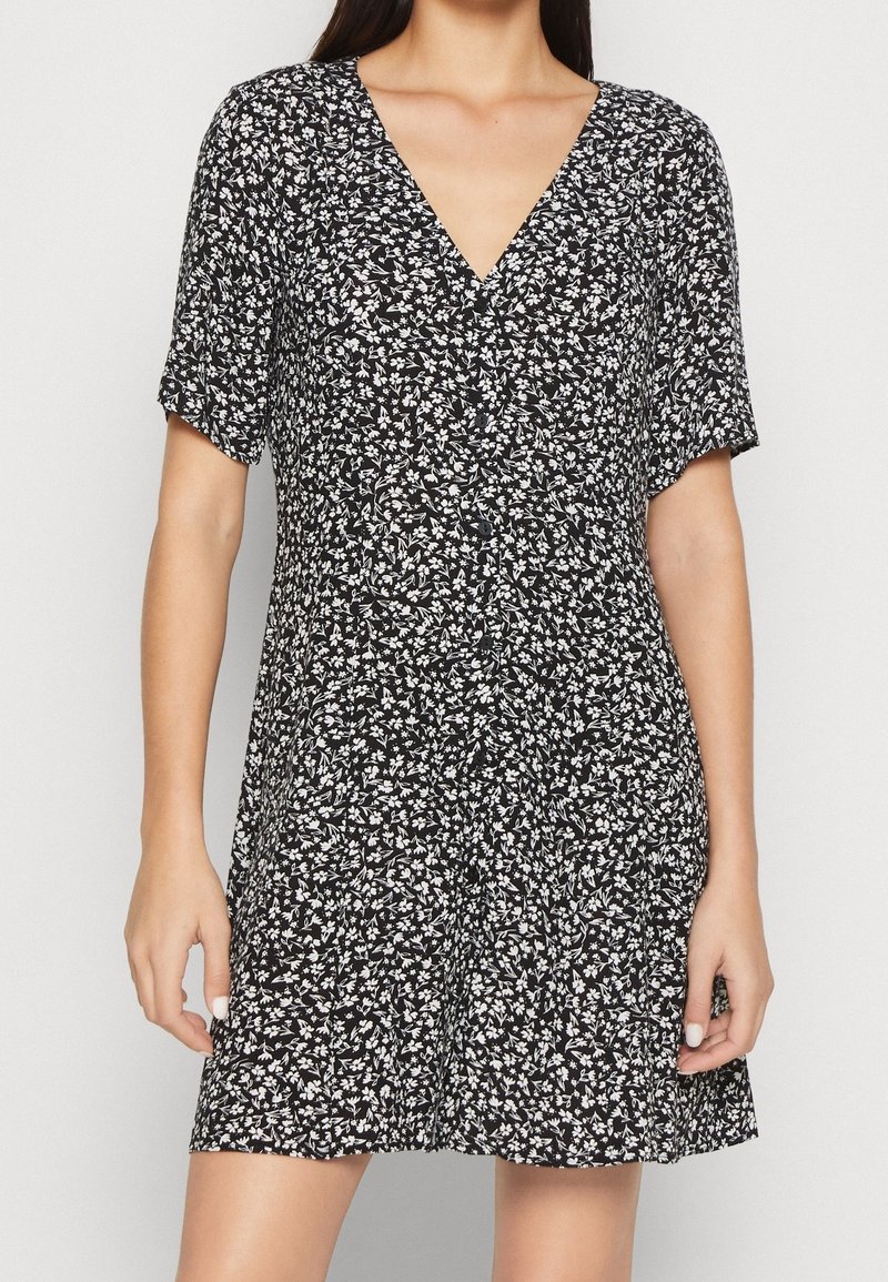 Femme portant une robe courte noire avec un imprimé floral blanc, col en V et manches courtes, debout devant un fond clair uni.