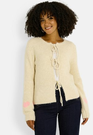 Cárdigan de punto color crema con acabado texturizado, con cierre frontal de lazo y detalles de corazones rosas en las mangas.