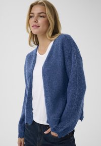 Blue knit cardigan made of soft, textured material. Features a relaxed fit, open front, and ribbed cuffs. Paired with a white shirt.