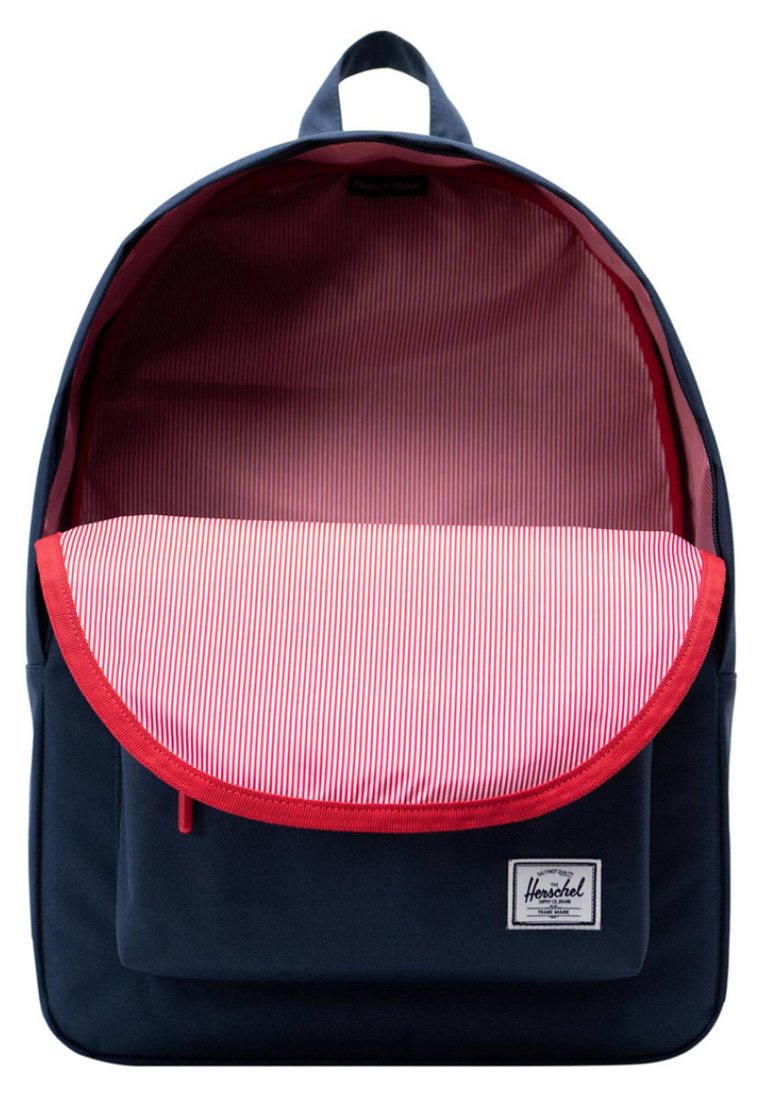 Un sac à dos bleu marine avec une doublure intérieure à rayures rouges et blanches, partiellement ouvert en haut, avec une poche avant et une étiquette de marque Herschel sur la poche avant.