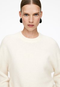 Cremefarbenes Sweatshirt mit einer weichen, gestrickten Textur und geripptem Rundhalsausschnitt. Akzentuiert mit großen schwarzen Ohrringen.