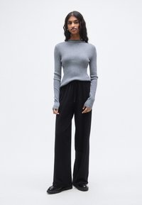 Γκρι μακρυμάνικη μπλούζα με ριμπ ύφανση και εφαρμοστό κορσάζ, σε συνδυασμό με φαρδιά μαύρη παντελόνα. Το μοντέλο φοράει μαύρα παπούτσια· σχεδίαση με μίνιμαλ αισθητική.