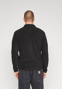 Chaqueta negra de forro polar con cuello, mangas largas y detalle de costura en la espalda. Se lleva con pantalones cargo oscuros que tienen un parche de logotipo rojo.