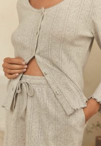 Person som bär en ljusgrå stickad cardigan och matchande drawstring-byxor, knäpper cardiganen med en hand, andra handen i fickan.