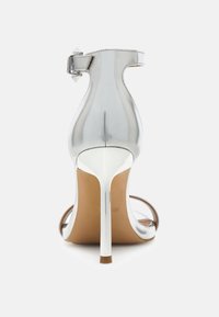 Zilveren sandaal met stilettohak, glanzende afwerking, enkelbandje en transparante zijkanten. Heeft een dunne, hoge hak en een beige zool.