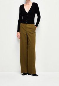 Donna che indossa un cardigan nero a scollo a V con volant, pantaloni a gamba larga color oliva e scarpe nere a punta, in piedi contro un muro bianco.