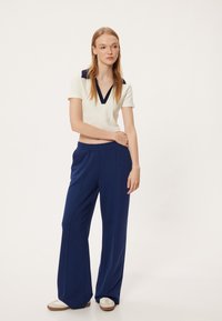 Witte top met korte mouwen en een marineblauwe kraag, gecombineerd met een losse marineblauwe broek. Het model draagt beige schoenen met witte accenten, tegen een effen achtergrond.