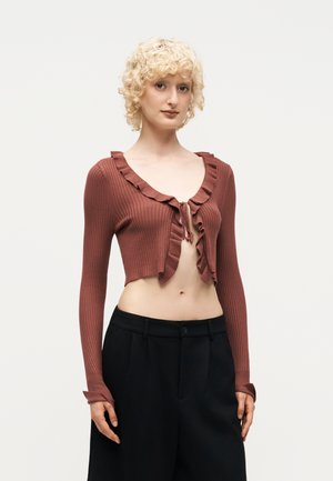 Blonde Frau, die eine kurz geschnittene, rostfarbene, gerippte Strickjacke mit Rüschen am Saum vorne gebunden und weite schwarze Hosen trägt.