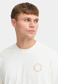 White cotton t-shirt with a round neckline and short sleeves. Features beige text in a circular pattern on the left chest area.