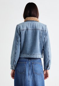 Džínová bunda se sherpa límcem, světle modré barvy, zkrácený střih, dlouhé rukávy. V kombinaci se stejným džínovým sukní.