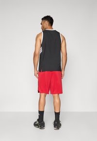 Mand, der står med ryggen til, iført sort ærmeløs sports T-shirt, røde træningsshorts, sorte ankelsokker og sorte sneakers med neonlys.