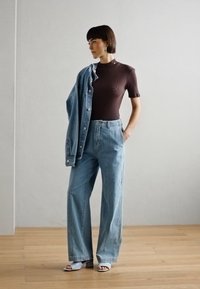 Braun gerippter, kurzärmeliger Oberteil, hellblaue, hochtaillierte, weit geschnittene Jeans, Jeansjacke über die Schulter gelegt und graue Sandalen auf einem Holzfußboden.