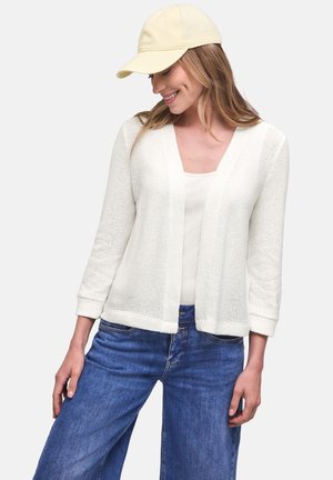 Kobieta nosząca kremowy, dziergany sweter na białym topie, niebieskie dżinsy i jasnżółtą czapkę z daszkiem, uśmiechająca się i patrząca w dół.